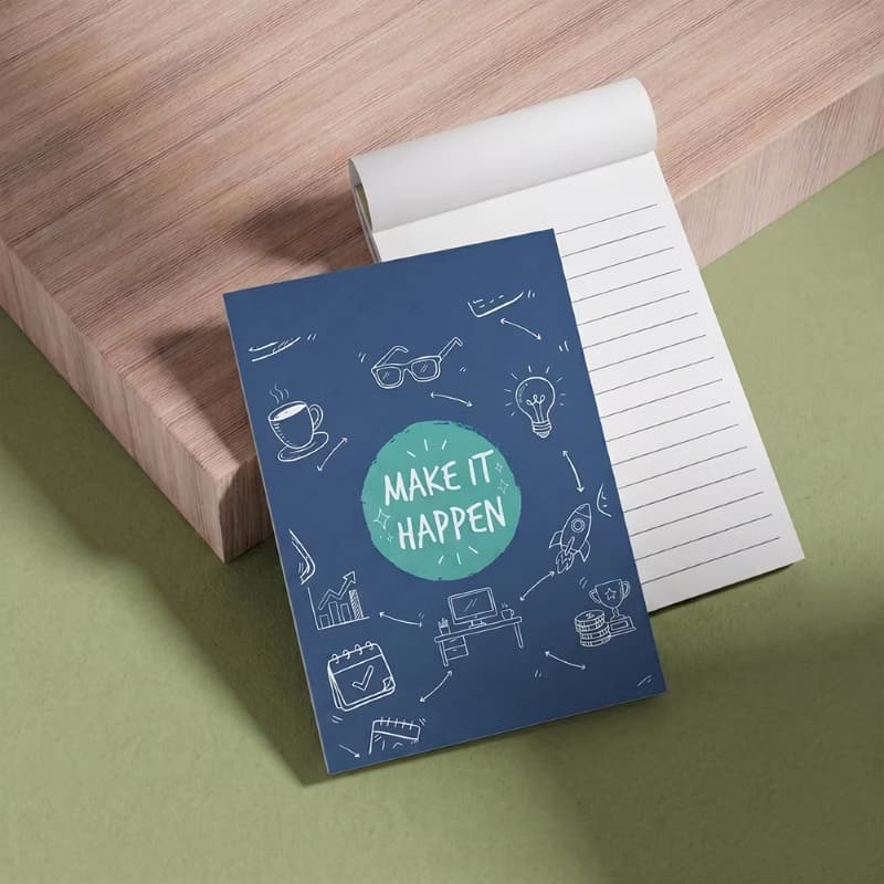 Custom Printed notepad with a blue "Make It Happen" motivational cover next to an open lined writing pad on a wooden block.