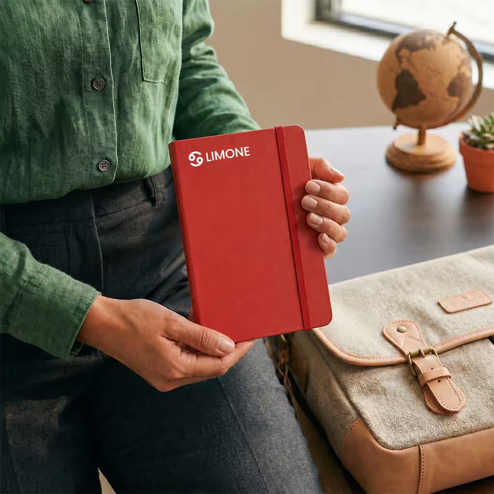 A person holding a red hardcover diary with an elastic band and a white "LIMONE" logo, sitting next to a messenger bag.