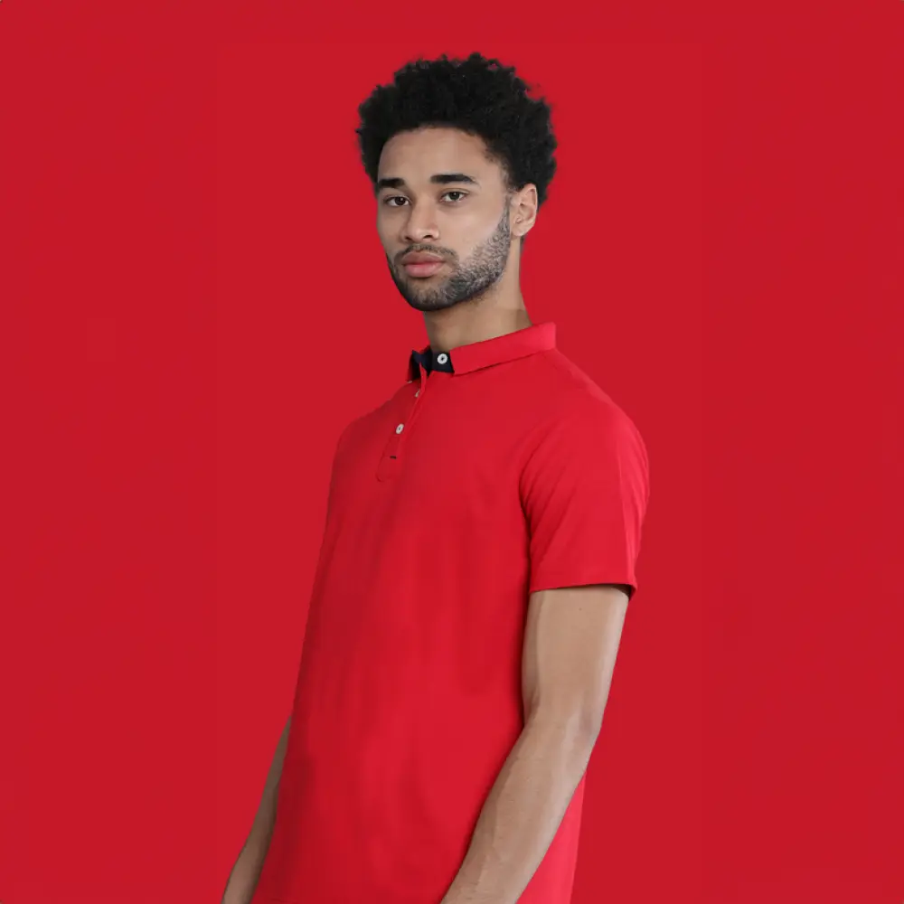 A man with dark curly hair and a beard wearing a vibrant red polo t-shirt with dark collar detailing, posing against a solid red background.