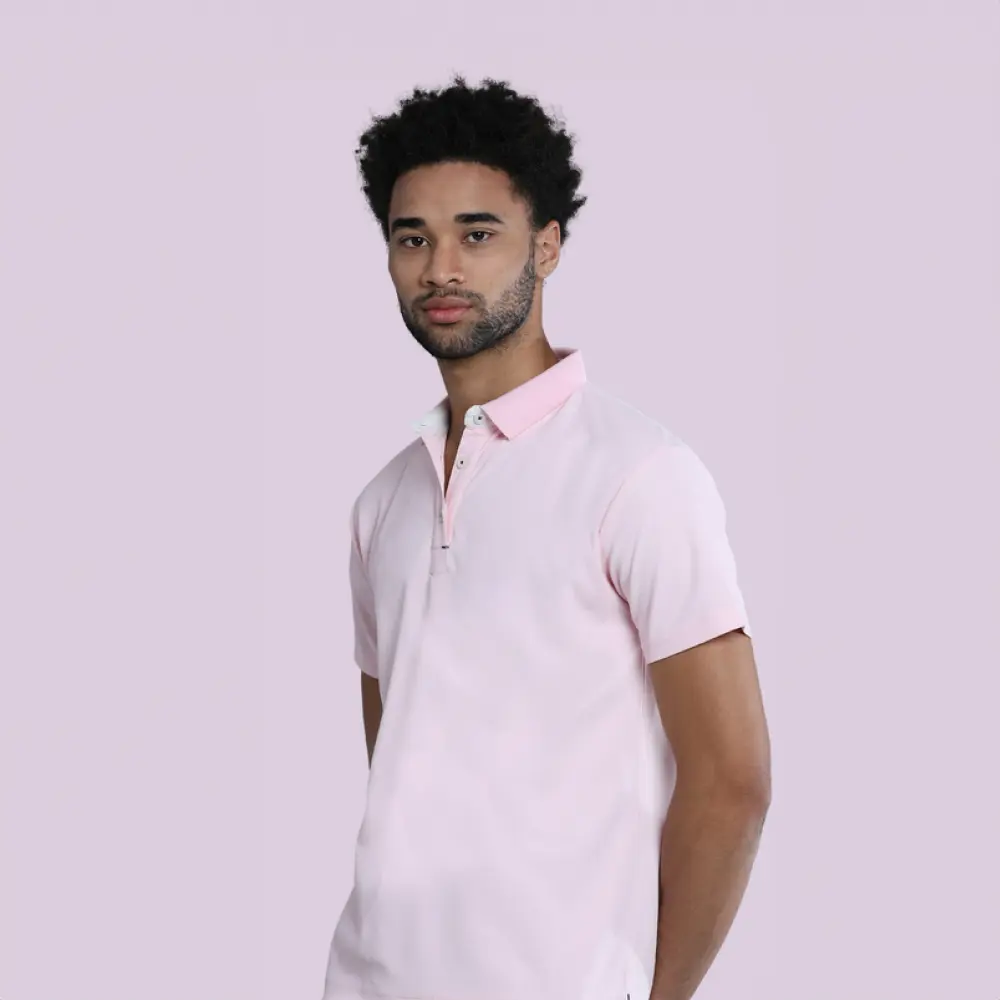 A man with curly hair wearing a light pink polo shirt, posing with his hands behind his back against a pale purple background