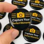 Black dome sticker with yellow camera icon, “Capture Your Perfect Moments,” close-up. Dome Sticker, custom sticker, 3D sticker.