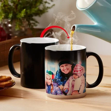 A sleek black magic mug with a personalized design revealed by hot liquid. The mug is shown on a wooden table with a steaming cup of coffee.