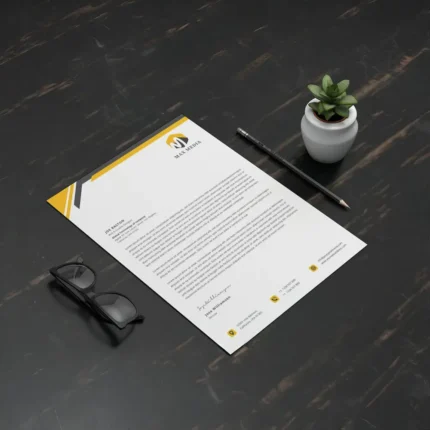 A custom letterhead with a yellow and black design, placed on a dark desk with glasses, a pencil, and a small plant.