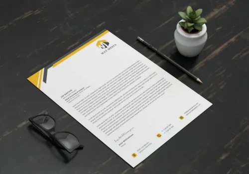 A custom letterhead with a yellow and black design, placed on a dark desk with glasses, a pencil, and a small plant.