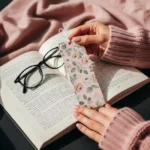 Hands with pink nails hold a custom floral Custom Bookmark over an open book in soft natural light.