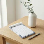Lined Notepads with a blue marble border and a silver pen on a light wood desk near a white vase.