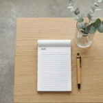 Minimalist lined Notepads with the word "NOTE" and a bamboo pen on a light wood table with a plant.