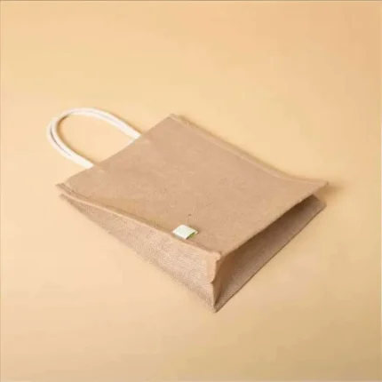 Angled top view of an empty, eco-friendly jute bag with white rope handles resting on a tan surface.