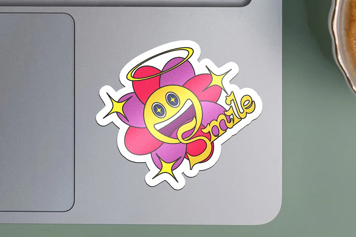 A die-cut sticker of a yellow smiling face with star-shaped eyes, surrounded by pink and purple flower petals, a halo above, and the word "Smile" in gold. The sticker is placed on a laptop.