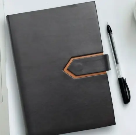 Black leather diary with a unique clasp design placed beside a black pen on a desk.