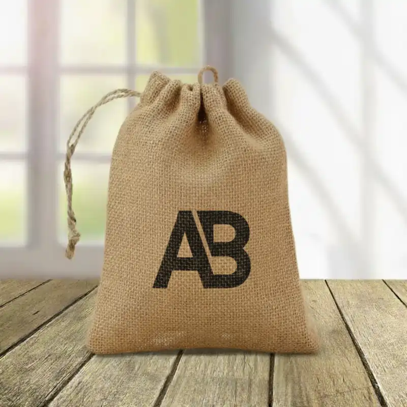 Drawstring Jute Bags with a custom black printed jute bag logo "AB" on a wooden table, featuring a natural, rustic look against a blurred window backdrop.