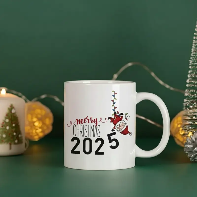 A white ceramic mug with "Merry Christmas 2025" and a cartoon Santa climbing Christmas lights, surrounded by festive decor.