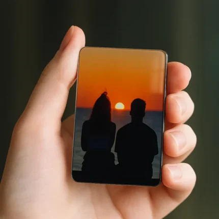 A hand holding a small rectangular magnet featuring a silhouette of a couple watching a sunset over the ocean