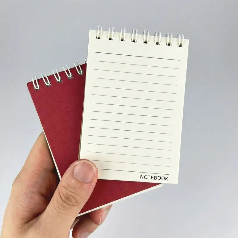 A hand holding two small spiral-bound pocket notebooks, one with a red cover and one open to a lined page.