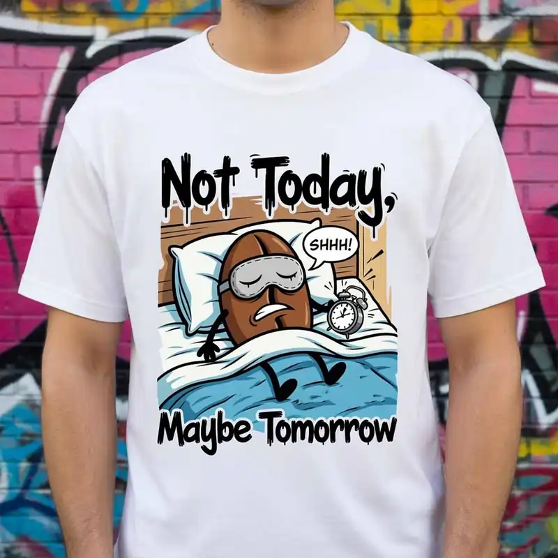 A white t-shirt featuring Funny Custom Print Ideas with a cartoon coffee bean in bed saying "Not Today, Maybe Tomorrow" to an alarm clock.