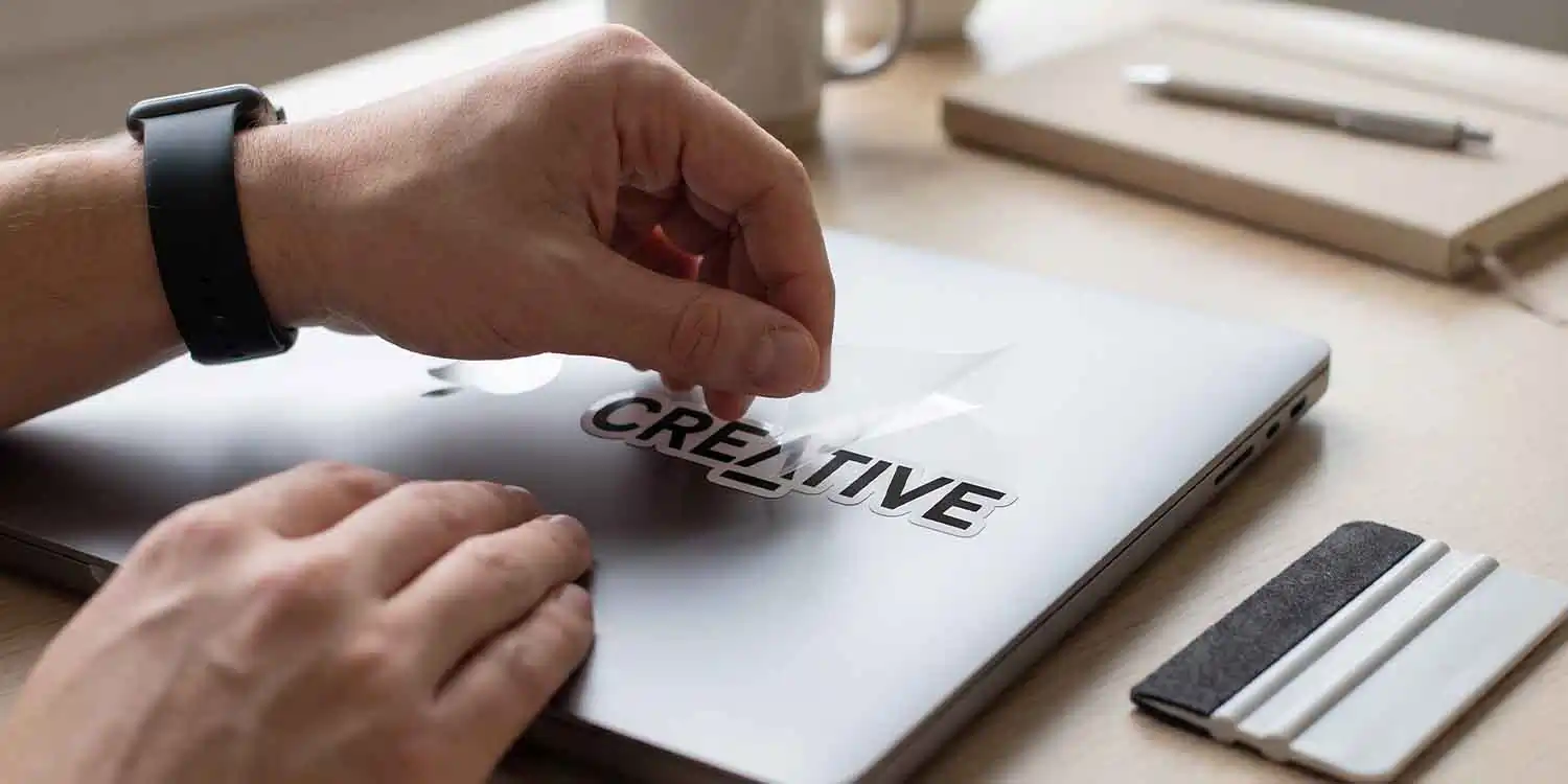 Hands applying a custom "CREATIVE" vinyl decal to a laptop using transfer tape for professional branding and commercial printing.